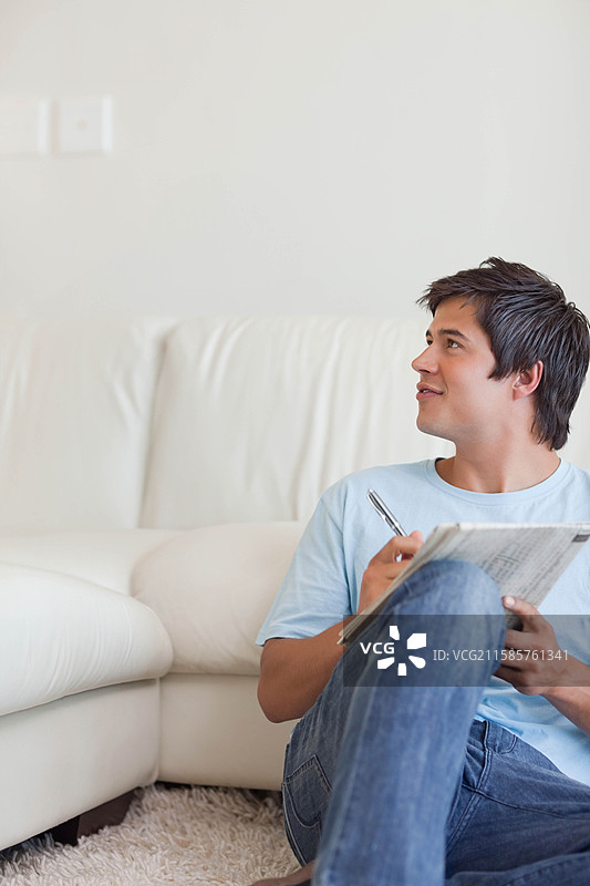 男人坐在客厅，靠在皮沙发上，手里拿着笔和报纸，留有空白空间。图片素材