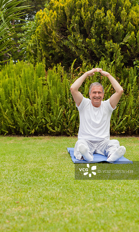穿着运动服的老年男子在后院的蓝色瑜伽垫上伸展双臂。图片素材