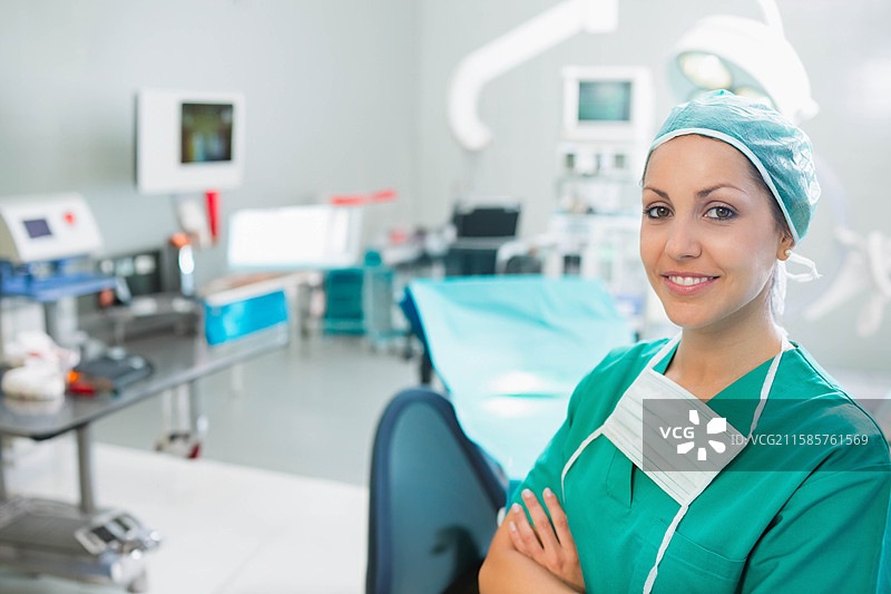 女性外科医生在手术室中双臂交叉，蓝色覆盖的手术台，灯光明亮，有空白区域可供使用。图片素材