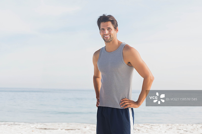 穿着灰色背心和海军蓝短裤的男子站在沙滩边缘，蓝天之下。图片素材