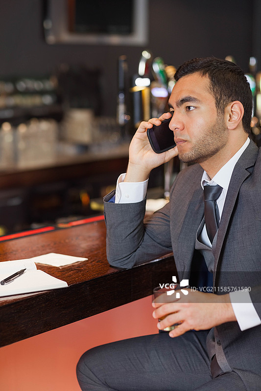 男性高管在木质酒吧台前一边喝琥珀色饮料一边用智能手机通话，旁边有记事本，留有空白空间。图片素材