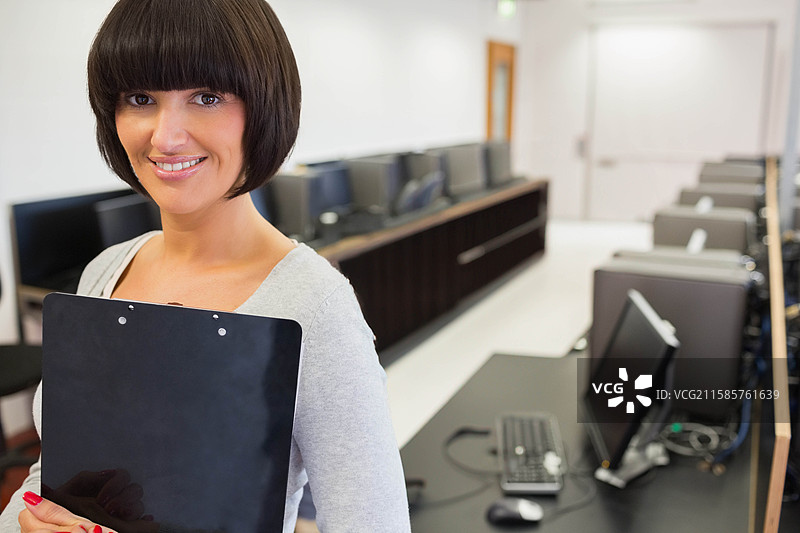 女性讲师站在计算机实验室左侧，手持夹板，靠近显示器，留有空白空间。图片素材