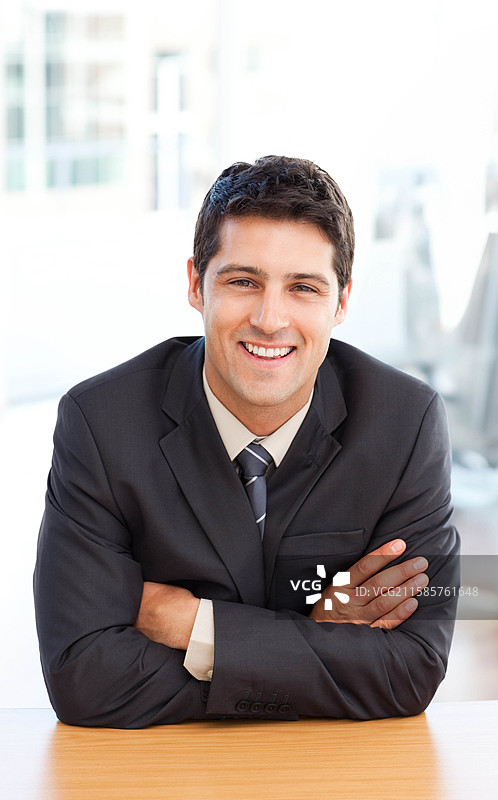 三十多岁的男性穿着西装和领带，坐在现代办公室的木桌前，面带微笑看向镜头。图片素材