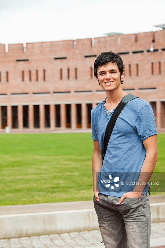 男大学生站在砖砌建筑旁的校园步道上，背着邮差包，留有空白空间。图片素材