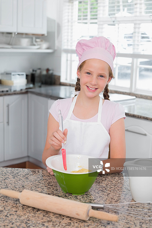 小女孩戴着粉色厨师帽和围裙，在家用厨房的搅拌碗中搅拌面糊。图片素材