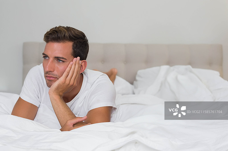 西班牙裔男子躺在卧室的白色床上，穿着白色T恤，靠近绗缝床头板。图片素材