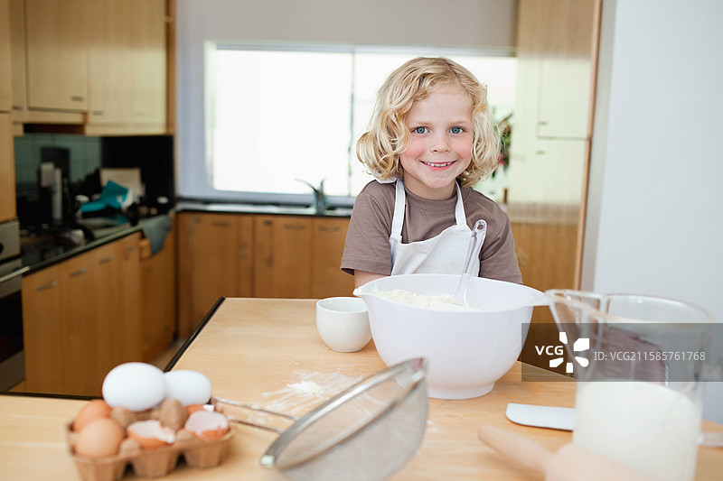 男孩穿着白色围裙，在厨房台面上搅拌食材，旁边有搅拌碗和鸡蛋 carton。图片素材