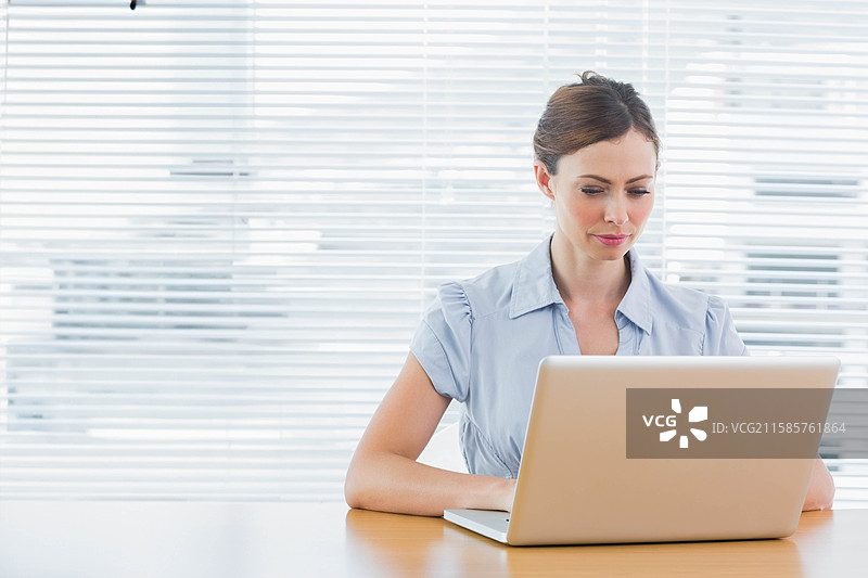 穿着蓝色衬衫的女性专业人士在办公室桌子上用银色笔记本电脑打字，窗帘旁有空白空间。图片素材