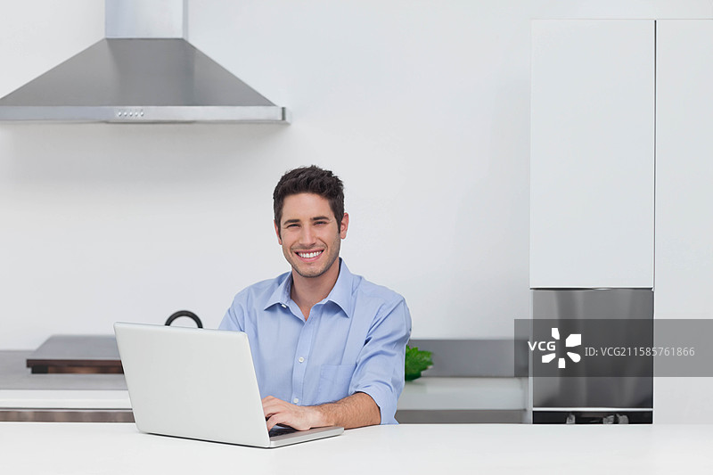 男子坐在现代厨房的白色台面上，使用银色笔记本电脑，旁边有一盆绿色植物。图片素材