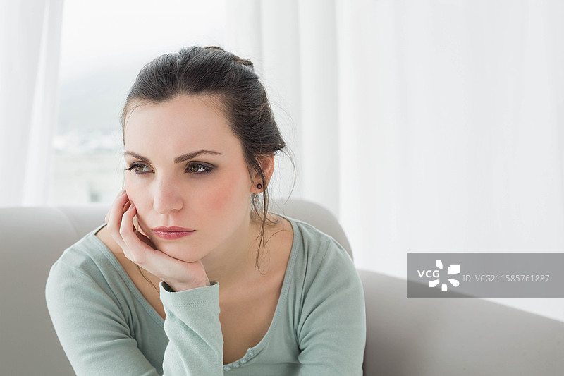 肤色白皙的女性坐在沙发上，透过客厅的白色薄纱窗帘凝视窗外。图片素材