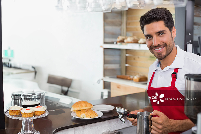 微笑的男咖啡师在柜台上手持咖啡粉斗和牛奶壶，旁边有杯子蛋糕，留有空白空间图片素材