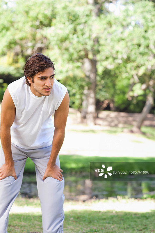 男子在公园草地上慢跑后停下，旁边是浅水池，穿着运动服，留有空白空间。图片素材