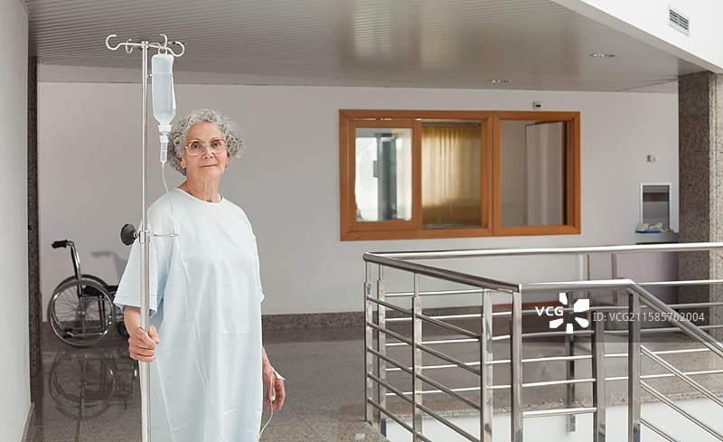 老年女性患者在病房走廊里扶着输液架，旁边有轮椅，留有空白空间。图片素材