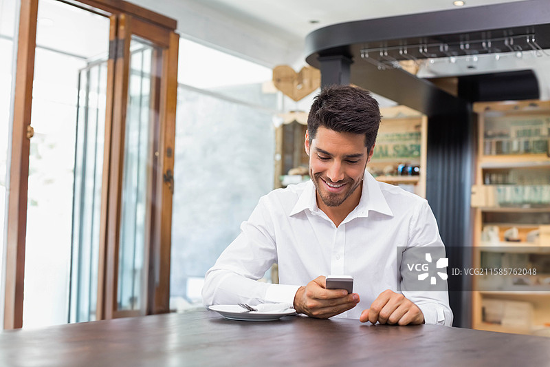 穿着白衬衫的男子坐在木制咖啡桌旁，查看智能手机，桌上有咖啡杯和盘子。图片素材