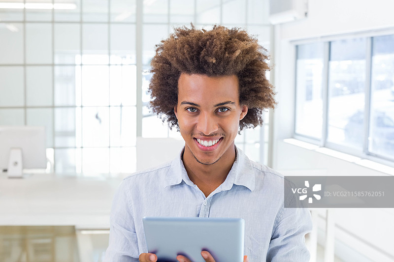 穿着浅蓝色衬衫的非裔美国男性在现代办公室里微笑着手持平板电脑。图片素材