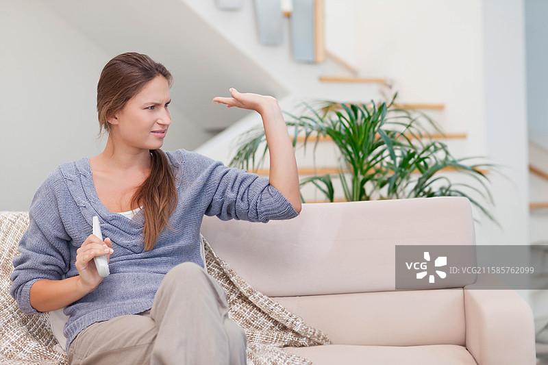 女人坐在米色沙发上，手持电视遥控器，抬起手掌，旁边是楼梯和一盆植物。图片素材