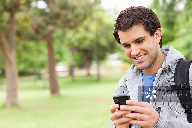 在郁郁葱葱的公园里，男人站着用智能手机轻敲，背着背包，留有空白空间。图片素材
