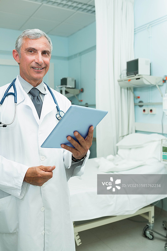 穿着实验室外套和听诊器的老年男性医生站在床边，手持平板电脑，留有空白空间图片素材