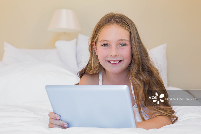 小女孩躺在卧室的床上，支着胳膊，手里拿着平板，周围是枕头和灯。图片素材