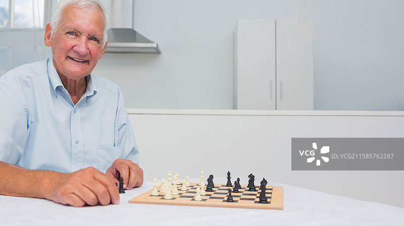 老年男子在厨房的木质棋盘上移动黑色棋子，白色橱柜背景，留有空白空间。图片素材