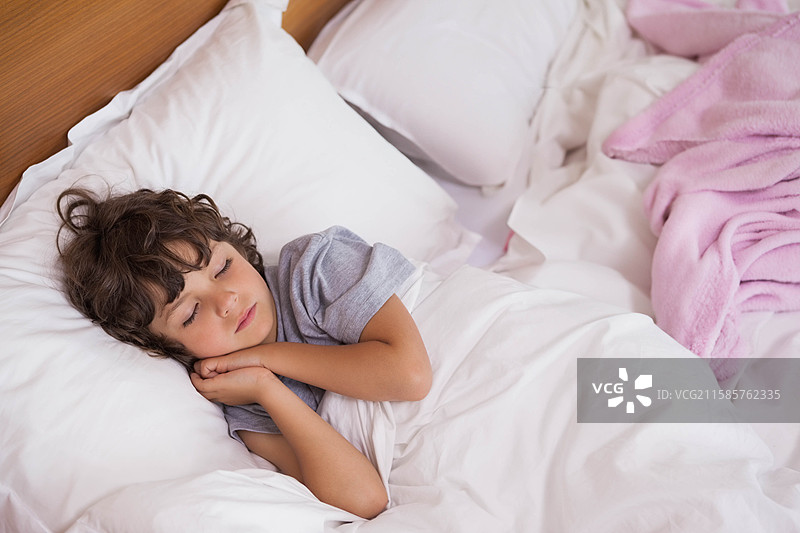 男孩侧卧在床上，白色床上用品、粉色毯子和木质床头板。图片素材