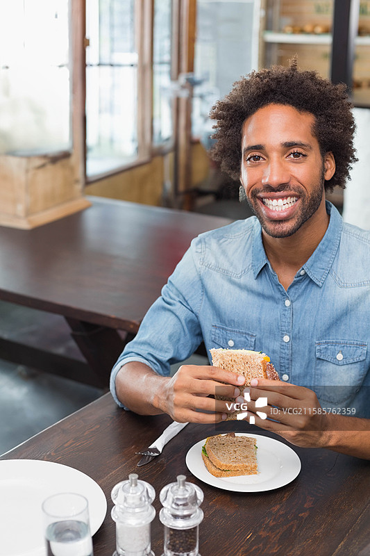 非裔美国男性坐在右侧的咖啡桌旁，手里拿着三明治，旁边有水杯，留有空白空间图片素材