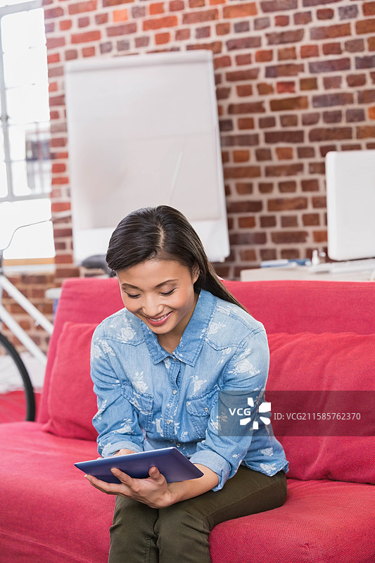 亚洲女性坐在红色沙发上，手持平板电脑阅读，周围是带有自行车轮的阁楼办公室休息区。图片素材