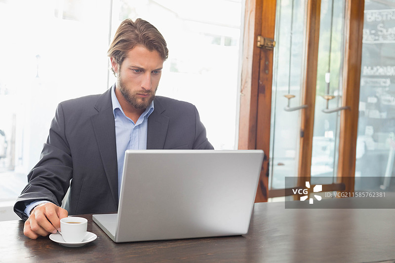 穿着西装的男子在窗边的咖啡店桌子上使用笔记本电脑，同时手里拿着咖啡，留有空白空间。图片素材