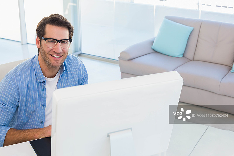 中年男子戴着眼镜，穿着衬衫，在家中的书桌上打字，显示出电脑显示器。图片素材