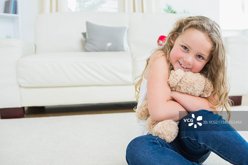 女童坐在地毯上，抱着泰迪熊，旁边是沙发垫，客厅里有空白空间。图片素材