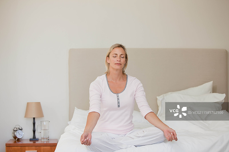 四十多岁的女性坐在卧室未整理的床上冥想，床头柜上有一盏台灯。图片素材