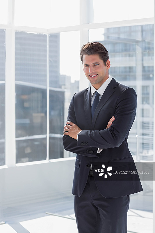 身穿商务西装的男性专业人士站在高层办公室的玻璃窗旁，双臂交叉。图片素材