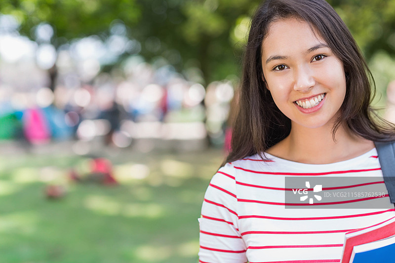 亚洲女学生站在校园广场上，手里拿着红色笔记本、蓝色文件夹和背包，留有空白空间。图片素材