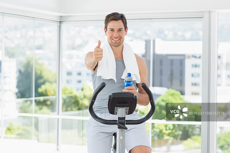 中年男子在健身房的窗边骑动感单车，手里拿着水瓶，头上搭着毛巾。图片素材