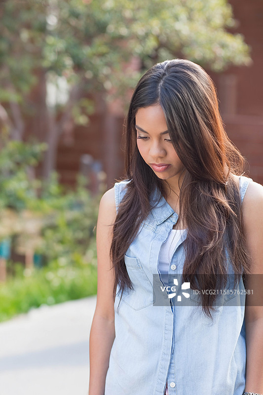 亚洲女性站在铺砌的花园小径上，穿着牛仔衬衫，低头看着绿色灌木。图片素材