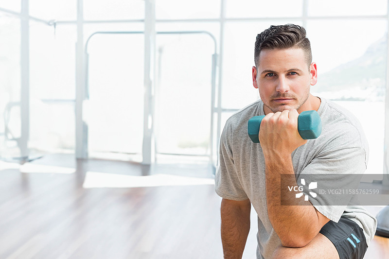 男子在健身工作室跪着，靠在玻璃墙旁用大腿弯举青绿色哑铃，留有空白空间。图片素材