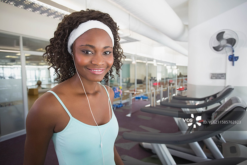 在健身房里，一位非裔美国女性站在跑步机旁，佩戴着汗带和耳机，留有空白空间。图片素材