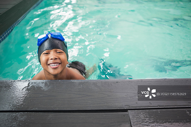 非裔美国男孩从泳池中走出，靠在木甲板上，戴着黑色帽子和蓝色泳镜。图片素材