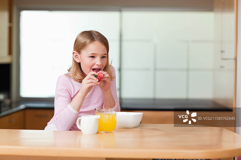 女孩在木质厨房台面上咬草莓，旁边有碗、果汁和杯子，留有空白空间。图片素材