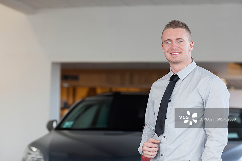 穿着衬衫和领带的成年男性在汽车展厅微笑，旁边是一辆深色轿车，留有空白空间。图片素材