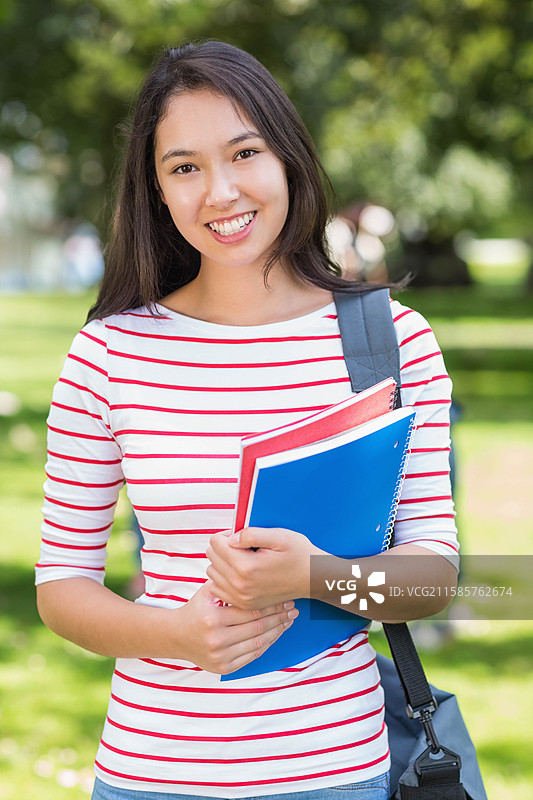 亚洲女学生站在校园草坪上，手里拿着红色和蓝色的螺旋笔记本，背着书包。图片素材