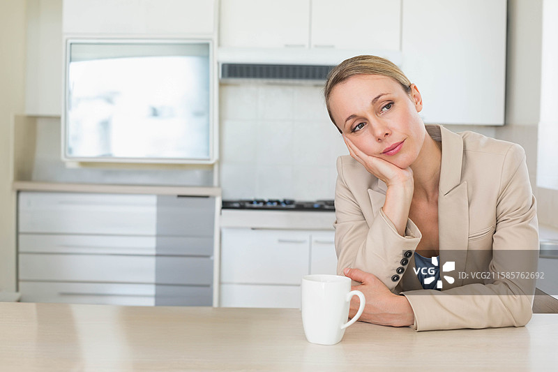 穿着米色西装外套的女性靠在厨房台面上，手肘支撑着，手里拿着咖啡杯，留有空白空间。图片素材