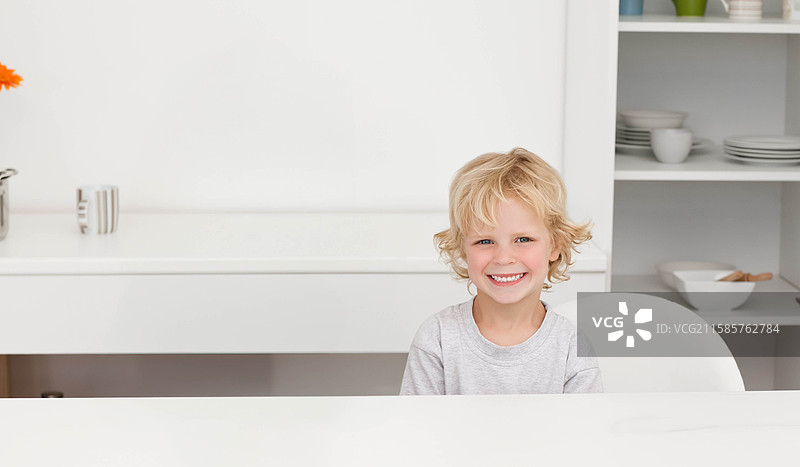 男孩坐在厨房的桌子旁，手里拿着杯子，旁边是花瓶和架子，留有空白空间。图片素材