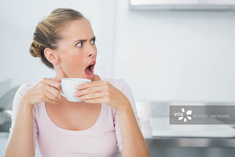 中年女性坐在厨房台面上，手里拿着白色杯子，面露震惊，留有空白空间图片素材