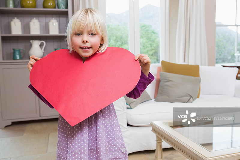 白人女孩站在客厅的灰色书架旁，手里拿着一个大红色纸心。图片素材