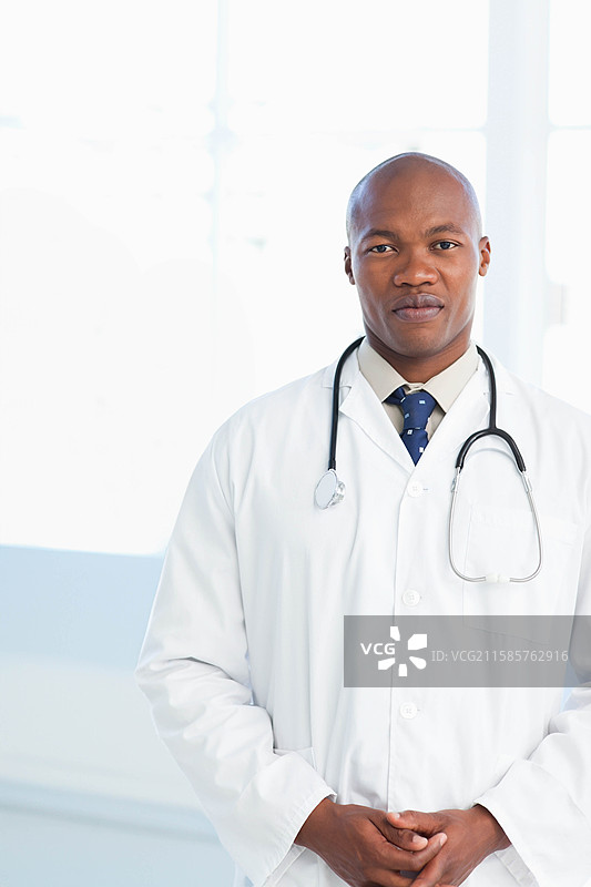 穿着白大褂、佩戴听诊器的非裔美国男性医生站在检查室内，留有空白空间图片素材