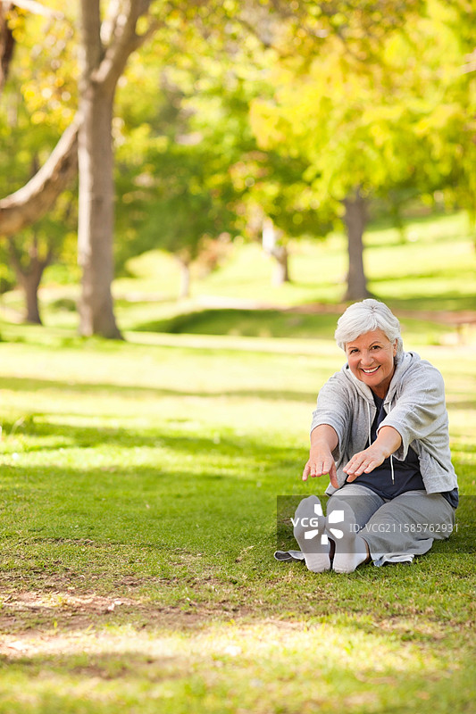 一位老年女性在阳光明媚的公园里坐在草地上伸展腿部并向前倾，留有空白空间。图片素材
