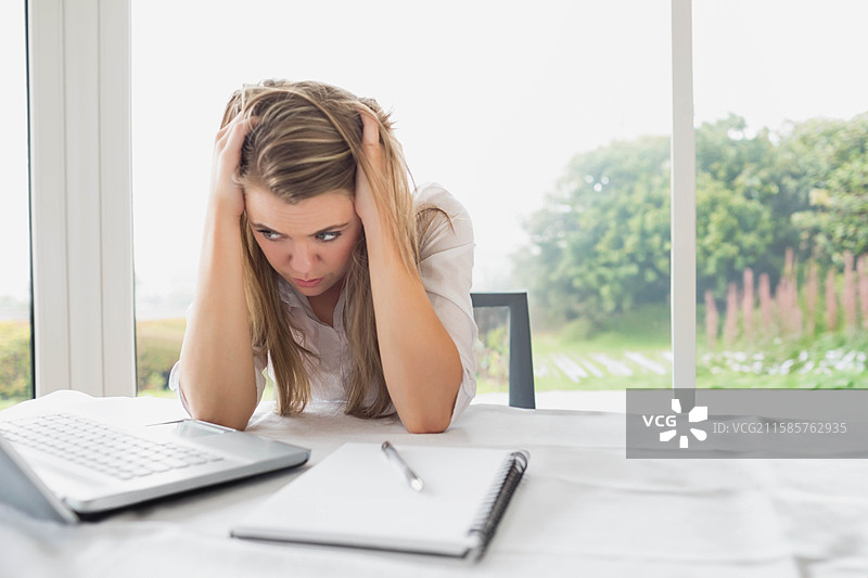 女人捧着头，凝视着窗边的桌子，桌上有笔记本、笔和一台笔记本电脑，窗外是花园景色。图片素材