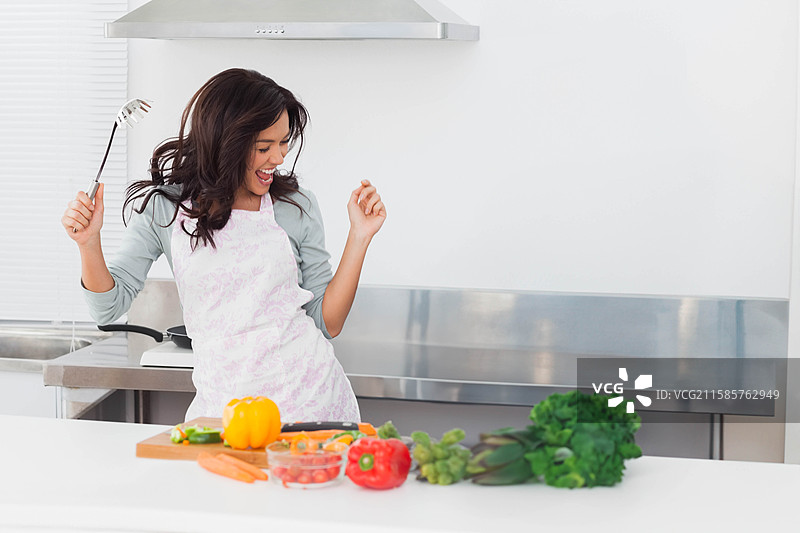 穿着淡色围裙的女人在厨房里跳舞，同时手里拿着金属勺，旁边是切好的彩椒。图片素材