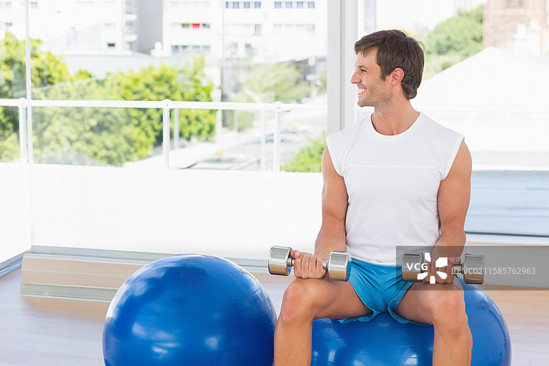 坐在稳定球上的男子手持哑铃，健身房内可俯瞰城市景观，留白空间。图片素材